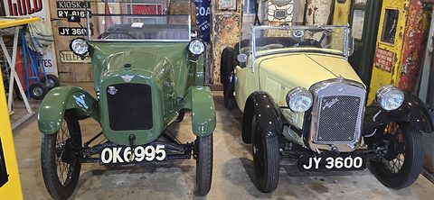 A green early open tourer and a cream later tourer side by side in a garage hung with signs.