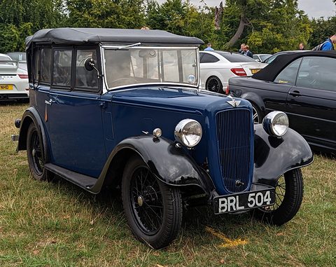A dark blue four-seat tourer with its hood up, a cowled grille and black wheels on grass. Opens a larger view
