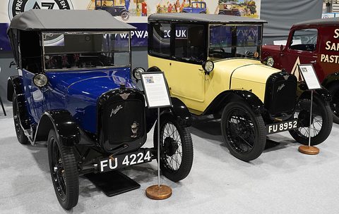 A blue open tourer with its hood up next to a yellow and black small saloon on a show stand. Opens a larger view