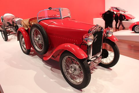 A bright red open two-seater with flowing wings and wire wheels on a white floor before a red wall. Opens a larger view