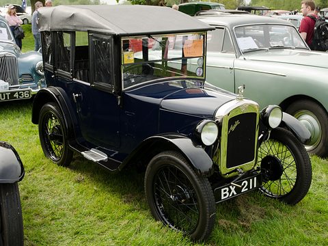 A dark blue open tourer with a grey hood up, black wings and wire wheels on grass beside other cars. Opens a larger view