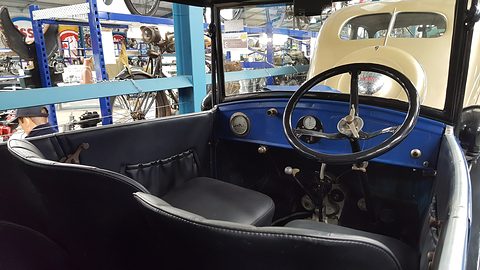 Looking into an open tourer with blue paint at a large black steering wheel, a small dashboard and black seats. Opens a larger view