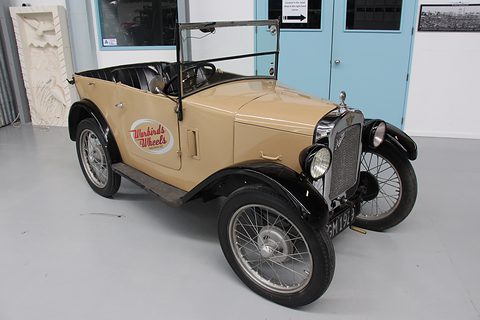 A small tan open tourer with black wings and wire wheels on a concrete floor in a hangar. Opens a larger view