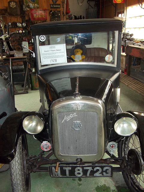 Head-on view of a dark small saloon with a nickel radiator in a crowded museum shed. Opens a larger view