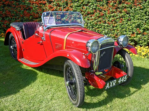 A red two-seater with sweeping wings, a chrome radiator and wire wheels on grass before a hedge. Opens a larger view