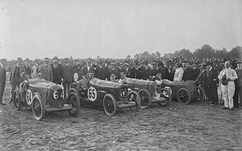 Black and white photograph of four small racing cars side by side in front of a large crowd. Opens a larger view