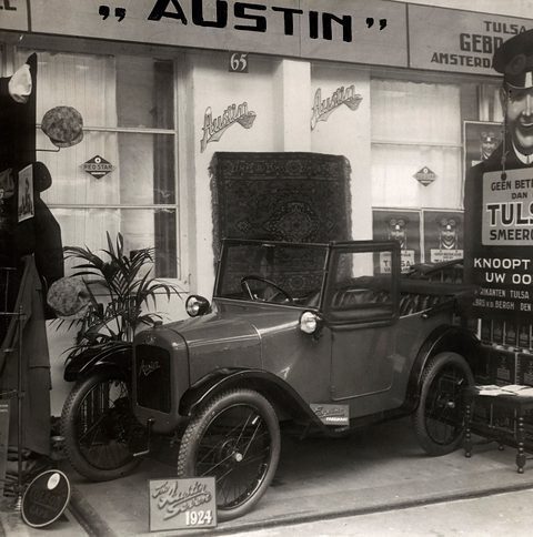 Black and white photograph of a small open tourer on a show stand under an Austin sign, a poster beside it.
