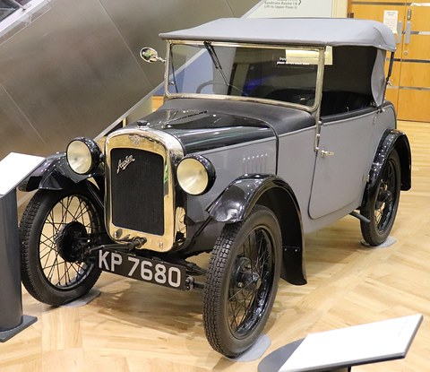 A grey two-seater with black wings and wheels and its hood up on a wooden museum floor. Opens a larger view