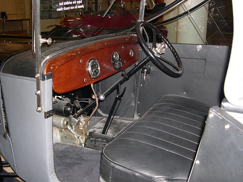 A wooden dashboard with round dials and a steering wheel in a grey-trimmed open cockpit. Opens a larger view