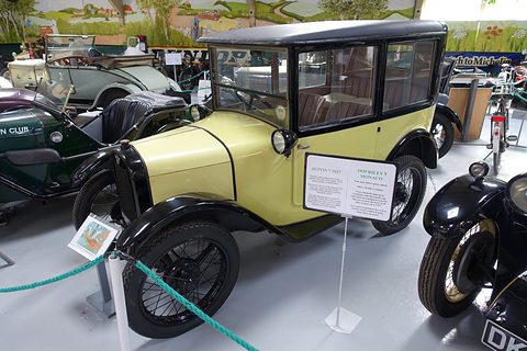 A pale yellow saloon with a black roof and wings behind a rope beside a display card. Opens a larger view