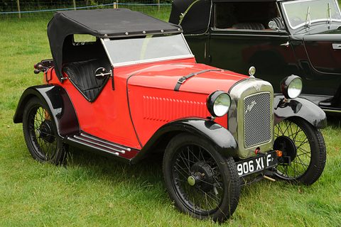 An orange two-seater with a black hood, black wings and wire wheels on grass. Opens a larger view