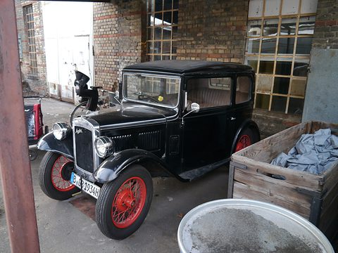 A black square saloon with red wheels parked in a cobbled yard beside a brick wall and crates. Opens a larger view
