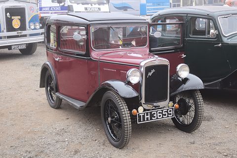 A maroon square-roofed saloon with black wings and wheels on a gravel yard beside other cars. Opens a larger view