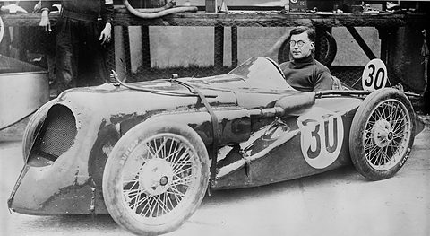 Black and white photograph of a bespectacled driver in a low streamlined racer numbered 30 in the pits. Opens a larger view