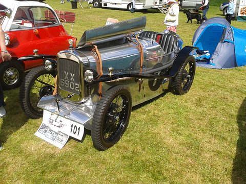 A polished aluminium two-seater with a long bonnet, wire wheels and a show board in front, on grass. Opens a larger view