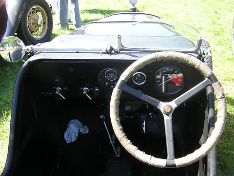 A black dashboard with dials behind a large cord-wrapped steering wheel in a small open car. Opens a larger view