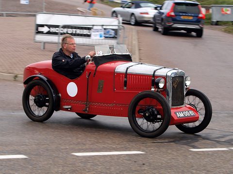 A red sports two-seater driven past a sign, its driver in a dark jacket. Opens a larger view