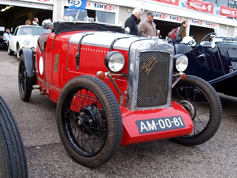 A red sports two-seater with a chrome radiator and wire wheels in a pit lane, people behind. Opens a larger view
