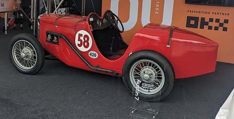 A red pointed-tail racing two-seater numbered 58 on a dark floor in front of a sponsor wall. Opens a larger view