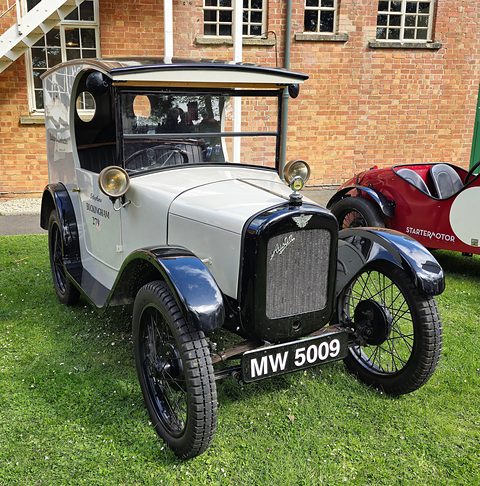 A cream van with an open-sided cab, black wings and wire wheels on grass by a brick building. Opens a larger view