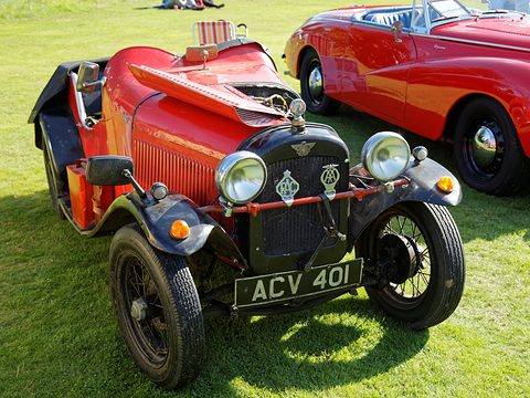A red open two-seater with a louvred bonnet strapped down and wire wheels on grass beside a red sports car. Opens a larger view