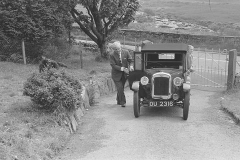 Black and white photograph of a small tourer with its hood up on a garden path, a man walking beside it. Opens a larger view
