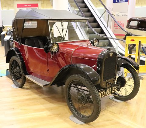 A dark red open tourer with a black hood up, black wings and wire wheels on a pale museum floor.