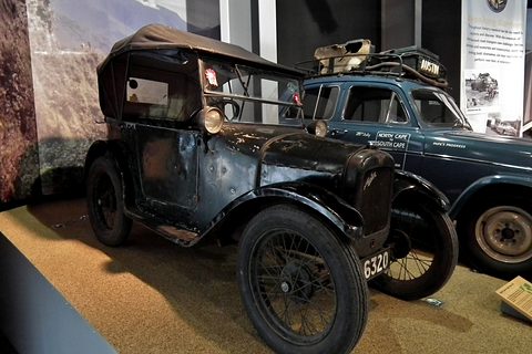 A worn black open tourer with its hood up and wire wheels in a dim museum display. Opens a larger view