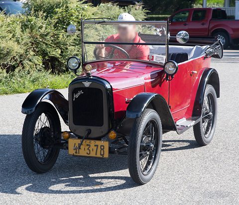 A small red open tourer with black wings, a black radiator and a yellow plate, its driver in the seat. Opens a larger view