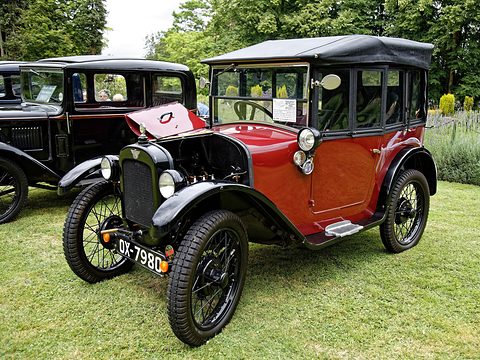 A red open tourer with a black hood up, black wings and wire wheels on a lawn by a hedge. Opens a larger view