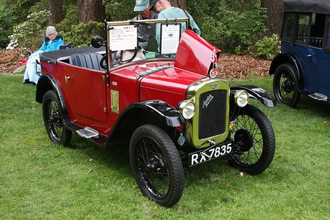 A red open tourer with black wings and its bonnet raised on grass, display cards on the windscreen. Opens a larger view