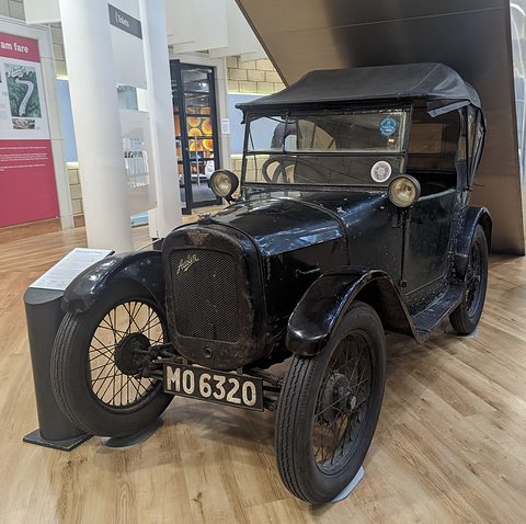 A small black open tourer with its hood up and wire wheels on a wooden museum floor beside a display stand. Opens a larger view