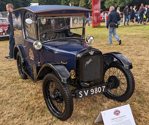 A small dark blue van with a gold-lettered panel side, wire wheels and black wings on grass. Opens a larger view