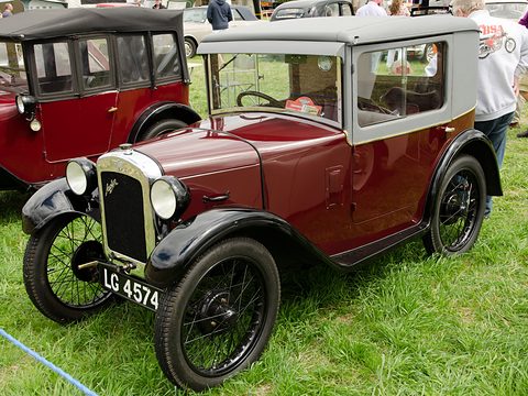 A maroon coupé with a fabric upper body, black wings and wire wheels on grass beside a red car. Opens a larger view