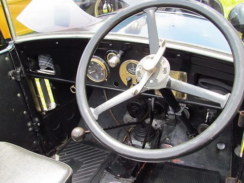 A large cream steering wheel in front of a black dashboard with round dials, pedals and levers below. Opens a larger view