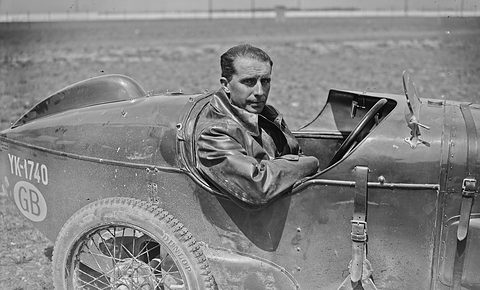 Black and white photograph of a driver in a leather coat sitting in a streamlined racer with GB and number plates.
