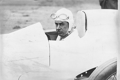 A man in a white helmet with goggles seated low in a smooth white streamlined body, a large wheel beside him. Opens a larger view