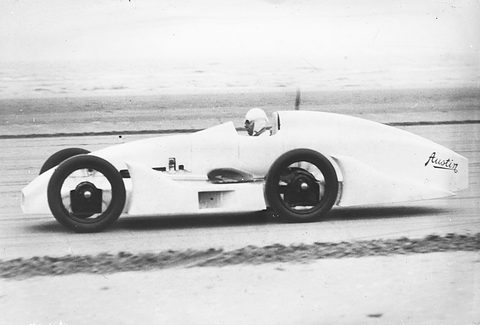 A long white streamlined single-seater with enclosed bodywork and exposed wheels racing across flat wet sand. Opens a larger view