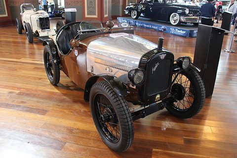 A small doorless sports two-seater with a polished aluminium bonnet and black wings on a wooden floor. Opens a larger view