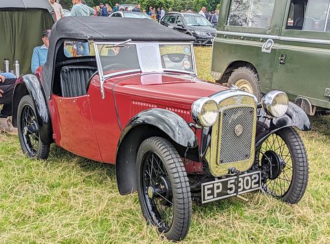 A red two-seater with a cowled radiator, black hood and wire wheels on grass, other cars behind. Opens a larger view