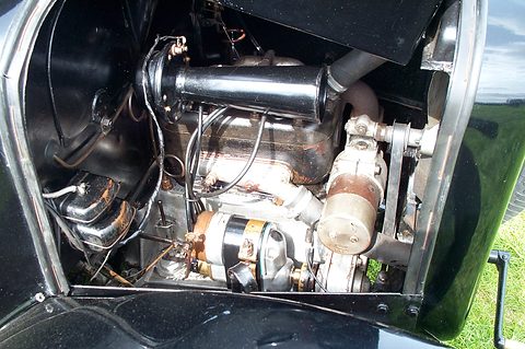 Close view of a small side-valve engine with a carburettor, exhaust pipe and spark plugs under a raised bonnet. Opens a larger view