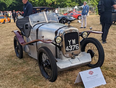 A silver pointed-tail two-seater with maroon wings and wire wheels on grass, a display card in front. Opens a larger view
