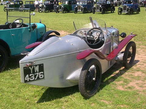 The rear quarter of a silver pointed-tail two-seater with maroon wings on grass among cars. Opens a larger view
