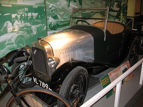 A polished aluminium two-seater with black wings and wire wheels before a painted racing mural. Opens a larger view