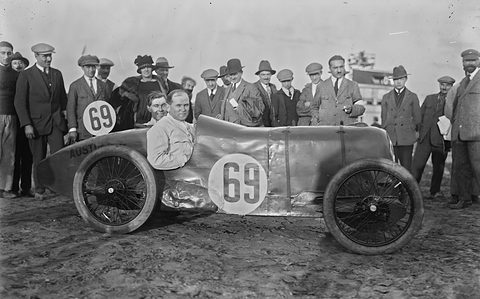 Black and white photograph of a dented streamlined racing car numbered 69, its driver smiling, onlookers behind. Opens a larger view