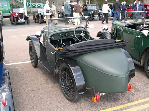 The rear quarter of a dark green pointed-tail two-seater in a paddock, people and cars behind. Opens a larger view