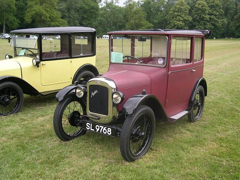 A maroon fabric-bodied small saloon with a sunshine roof and wire wheels on grass beside a cream car. Opens a larger view