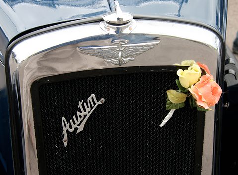 A chrome radiator shell with a winged mascot above and the word Austin in script on the grille, flowers beside it. Opens a larger view