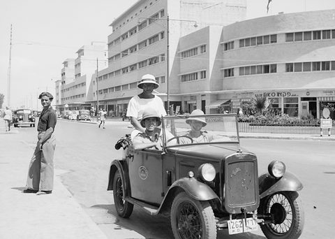 Black and white photograph of a small open tourer with uniformed men aboard on a wide street of modern buildings.