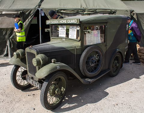 A camouflaged green van with Home Guard lettering parked on gravel by army tents. Opens a larger view